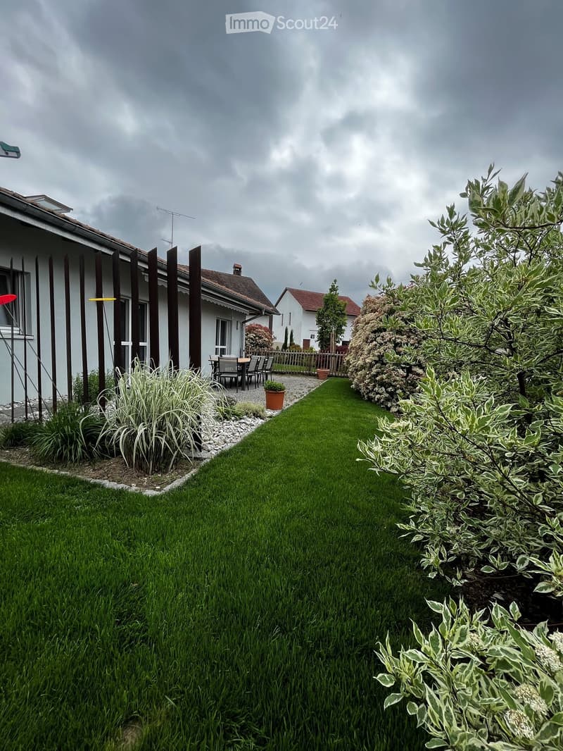 Well-maintained garden with a lawn, various plants, and a patio area with a table and chairs near the house.