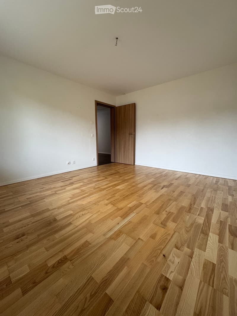 Empty room with light-colored wooden flooring and white walls, featuring a single open wooden door leading to another space.