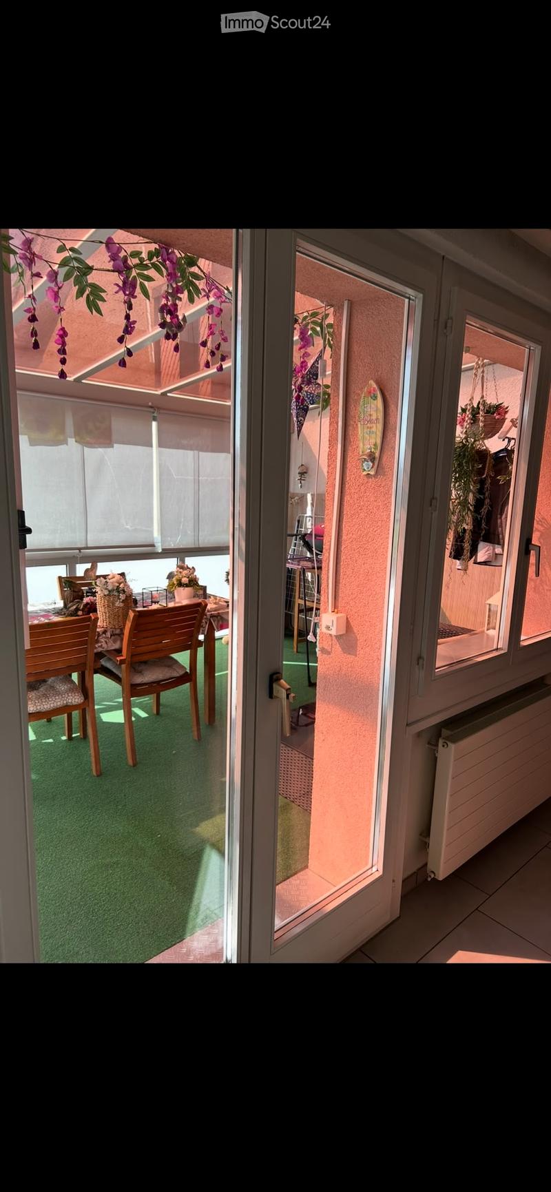 Balcony with green artificial turf, wooden chairs, small table, and flower pots. The balcony has a glass door leading to the interior, with hanging plants and a surfboard decoration visible.