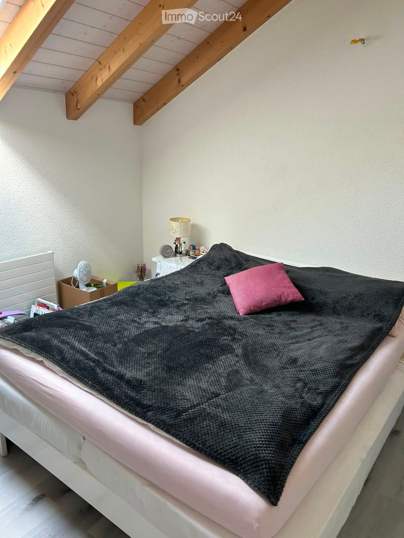A cozy bedroom with a neatly made bed, a nightstand with a lamp, and a fan in the corner.