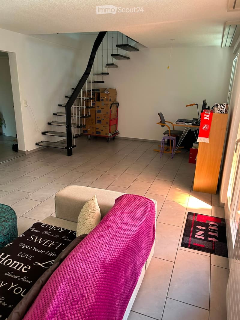 A furnished living room with a sofa, a pink blanket, a desk with a laptop, a chair, and a staircase leading to another level.