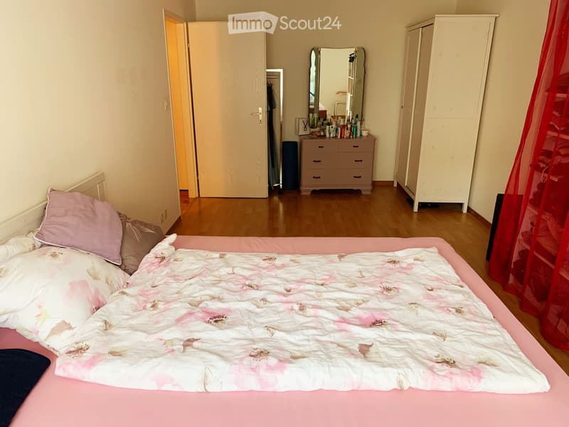 Bedroom with a pink bed, floral duvet, pillows, a large mirror, a wardrobe, and a dresser with various items on top.