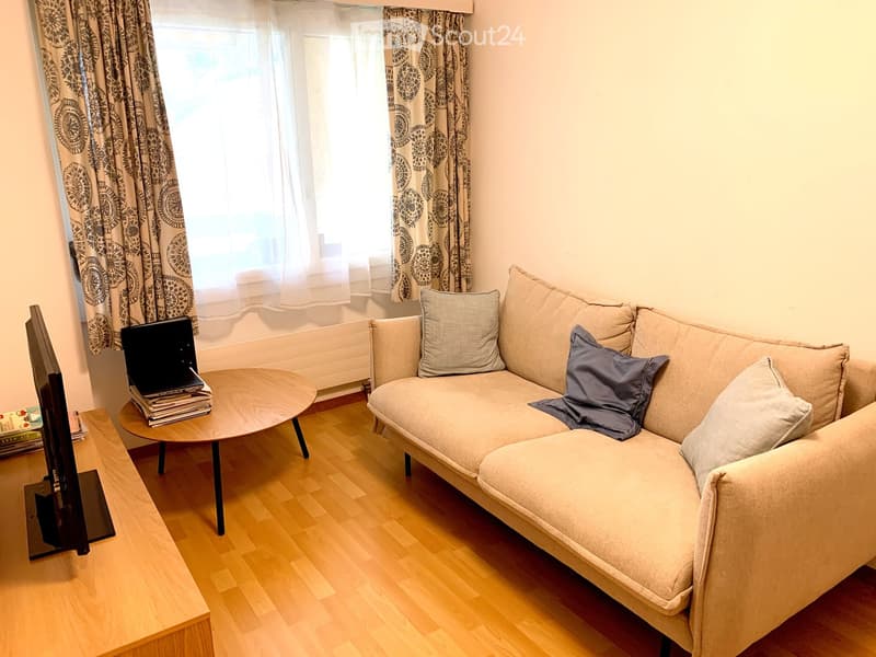 Living room with a light-colored sectional sofa, wooden coffee table, laptop, and television. The room features wooden flooring and large windows with patterned curtains.