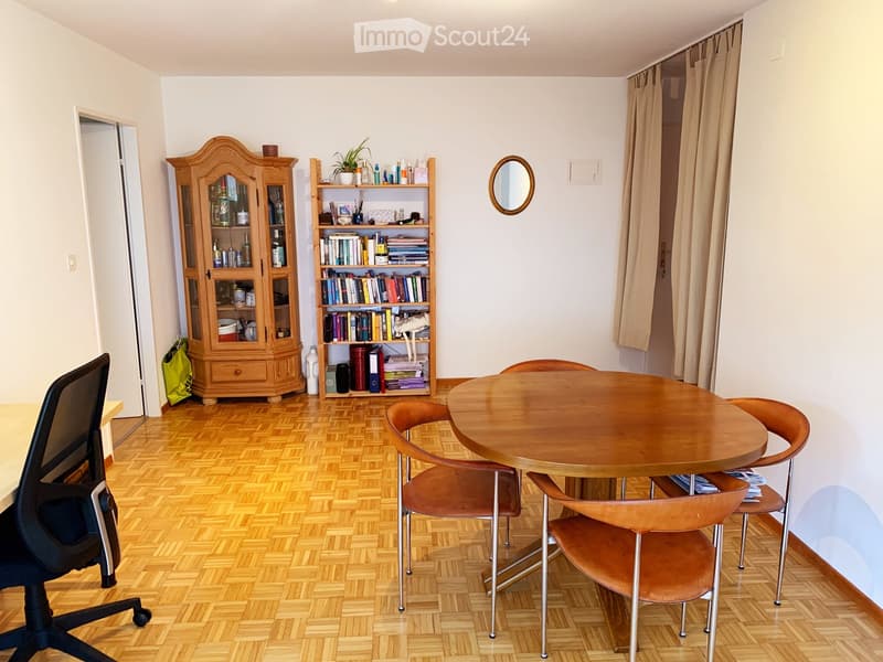 A well-lit room with wooden flooring, a round wooden table with four chairs, a wooden cabinet, a bookshelf filled with books and decorative items, and a small round mirror on the wall.