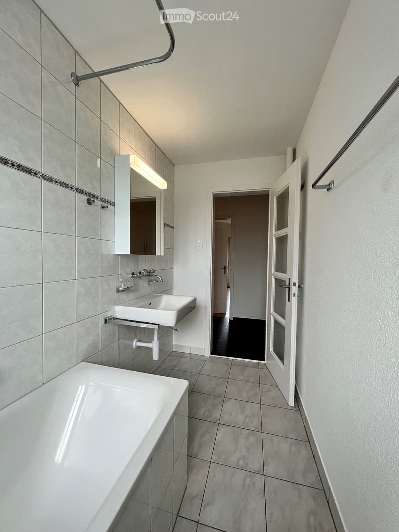 Modern bathroom with a white bathtub, sink, and mirror. The walls are tiled in grey, and the floor is tiled in grey as well. There is an open door leading to another room.