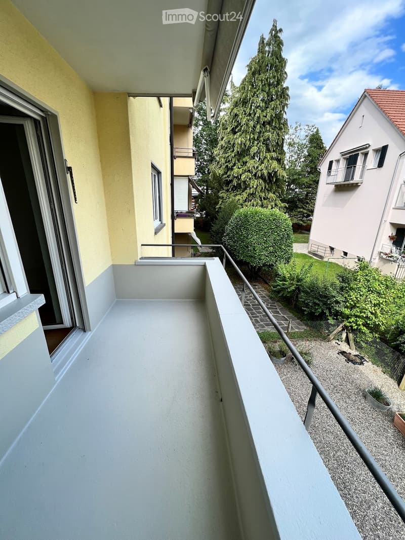 Balcony with white concrete floor, metal railing, and sliding glass door leading to the interior. The view includes a garden area with bushes, a tree, and neighboring buildings.