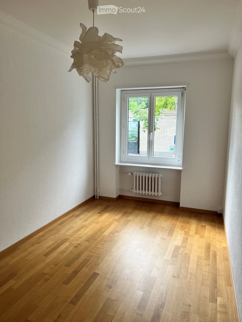 Empty room with light wooden flooring, white walls, a large window with white frame, radiator below the window, and a modern hanging light fixture.