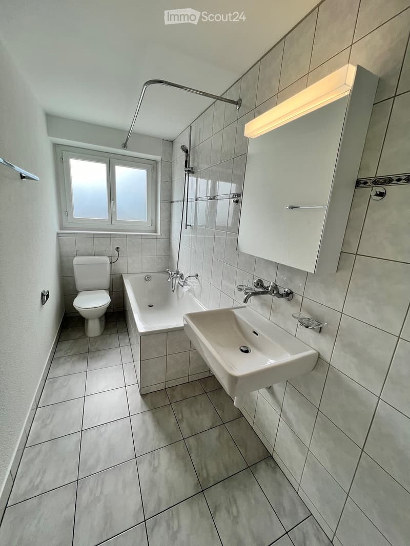 Modern bathroom with white tiles, a toilet, bathtub, sink, and a shower area with a glass enclosure. Features a window with frosted glass panes and built-in cabinets.