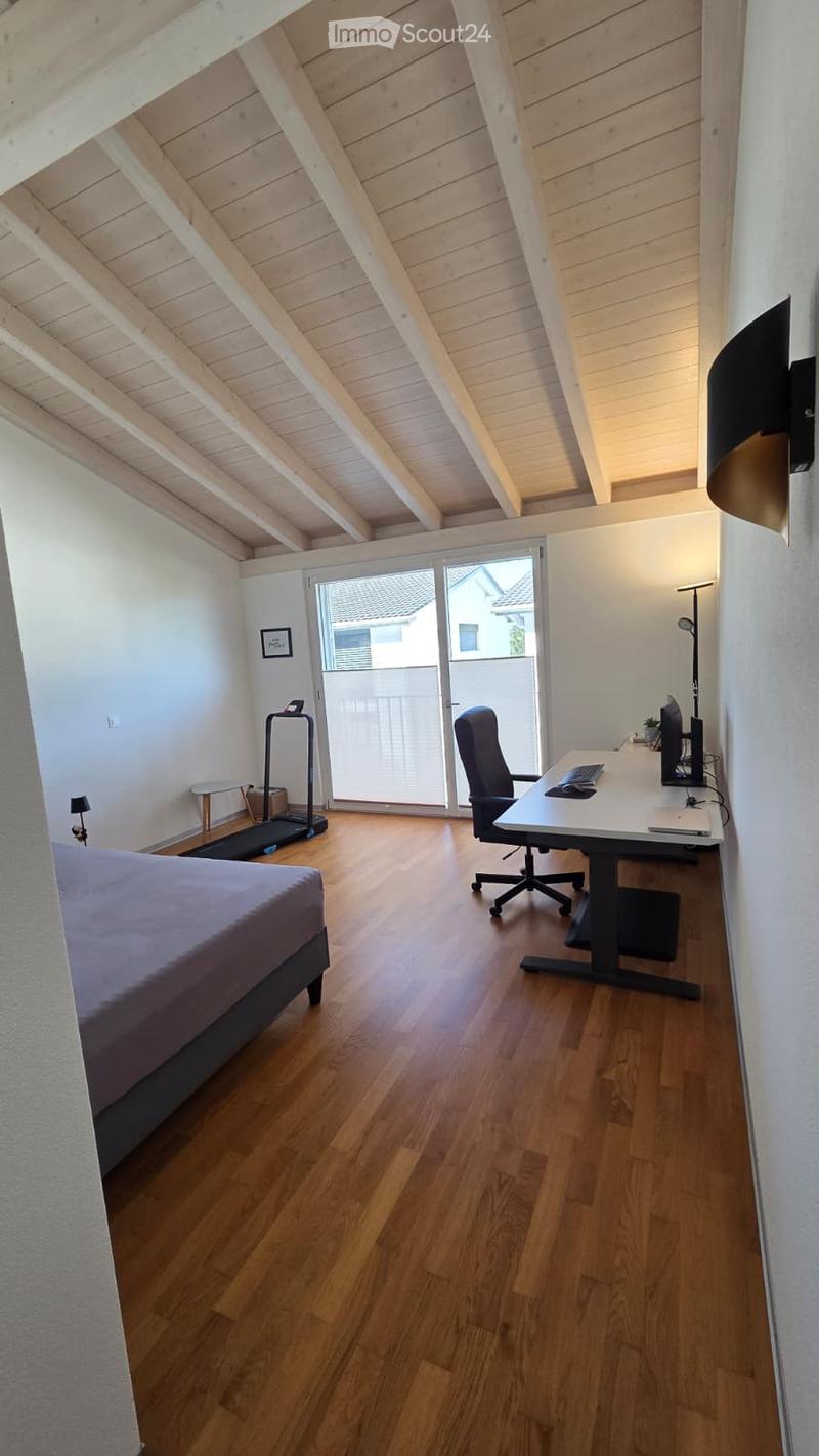 A multi-functional room with a bed, desk, and exercise bike. The room features wooden flooring, white walls, and large windows with a view of the outside.