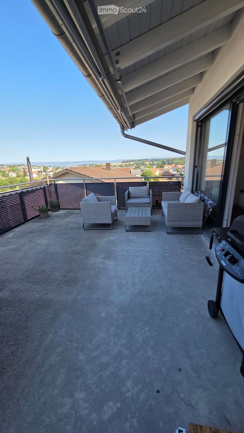 A modern balcony with a concrete floor, featuring a sofa, chairs, a small table, and potted plants, with a scenic view of the town and countryside.