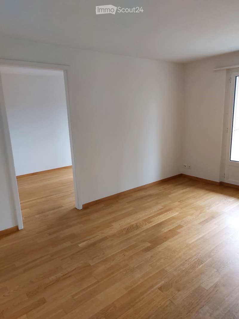 Empty room with light wooden flooring and white walls, featuring a doorway and a window.