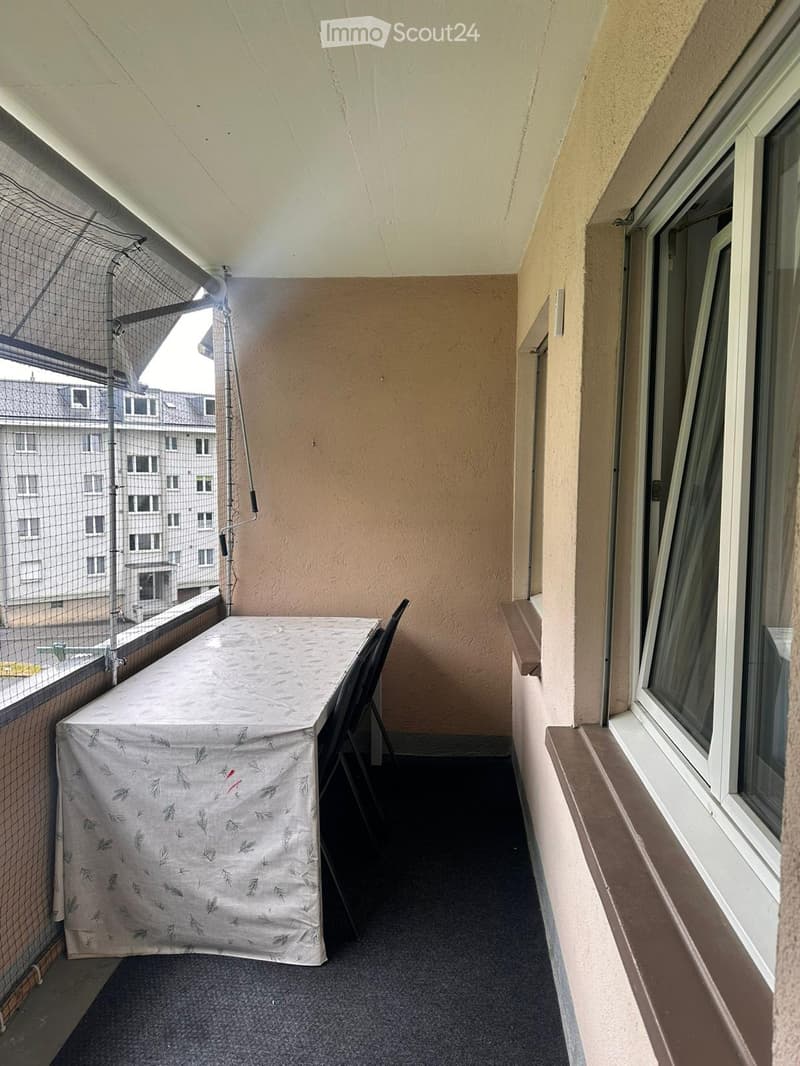 Balcony with a covered table and a chair, overlooking a cityscape with buildings.
