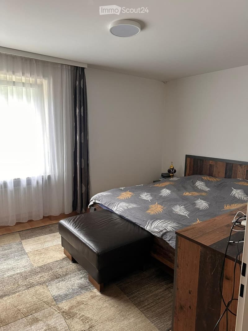 Modern bedroom with a wooden bed frame, grey bedding with leaf patterns, a black leather stool, a wooden nightstand, and a lamp.