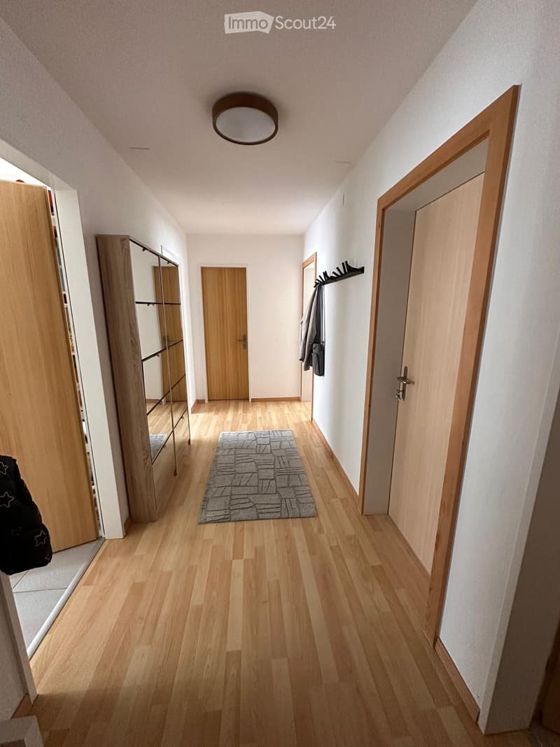 A narrow hallway with wooden flooring, white walls, and several doors leading to different rooms. A small rug is placed in the center, and a coat rack with jackets is visible on the right side.