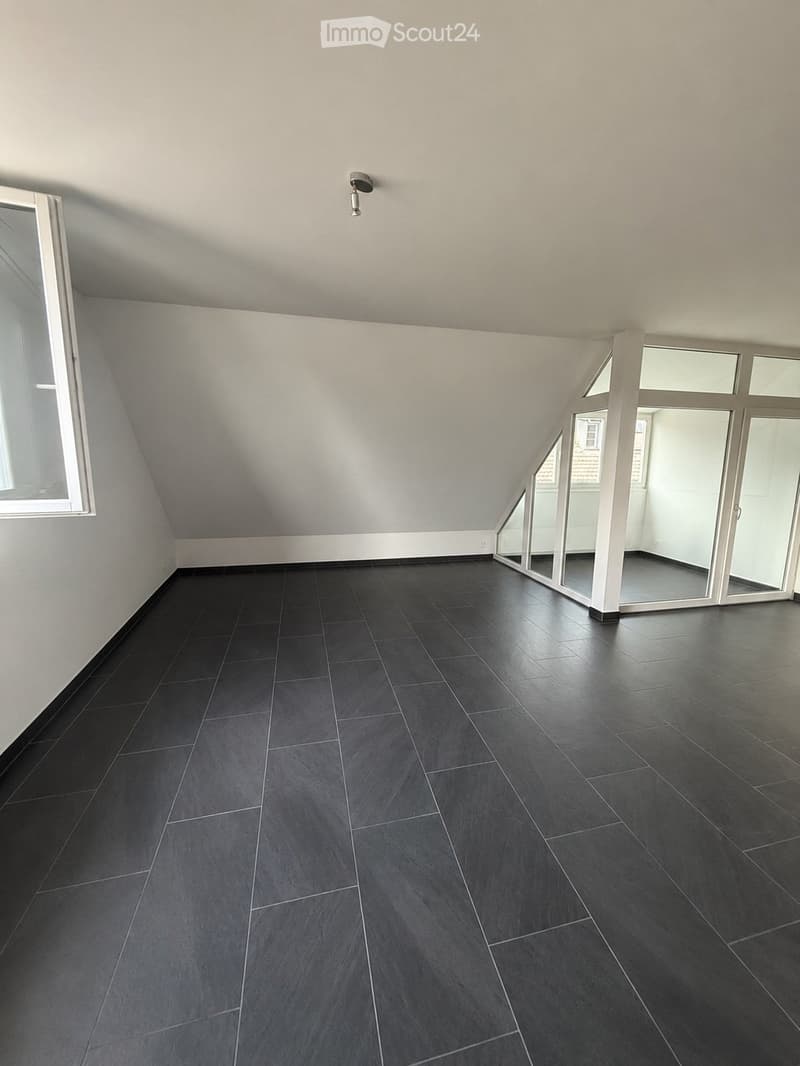 Empty room with dark grey tiled floor and white walls, featuring large windows and a sliding glass door leading to another space.
