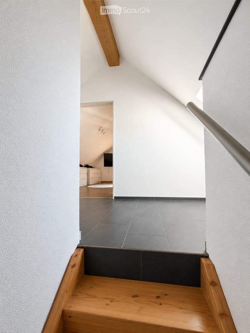 Modern staircase with wooden steps, metal railing, and grey tiled landing leading to an open area with white walls and wooden flooring.