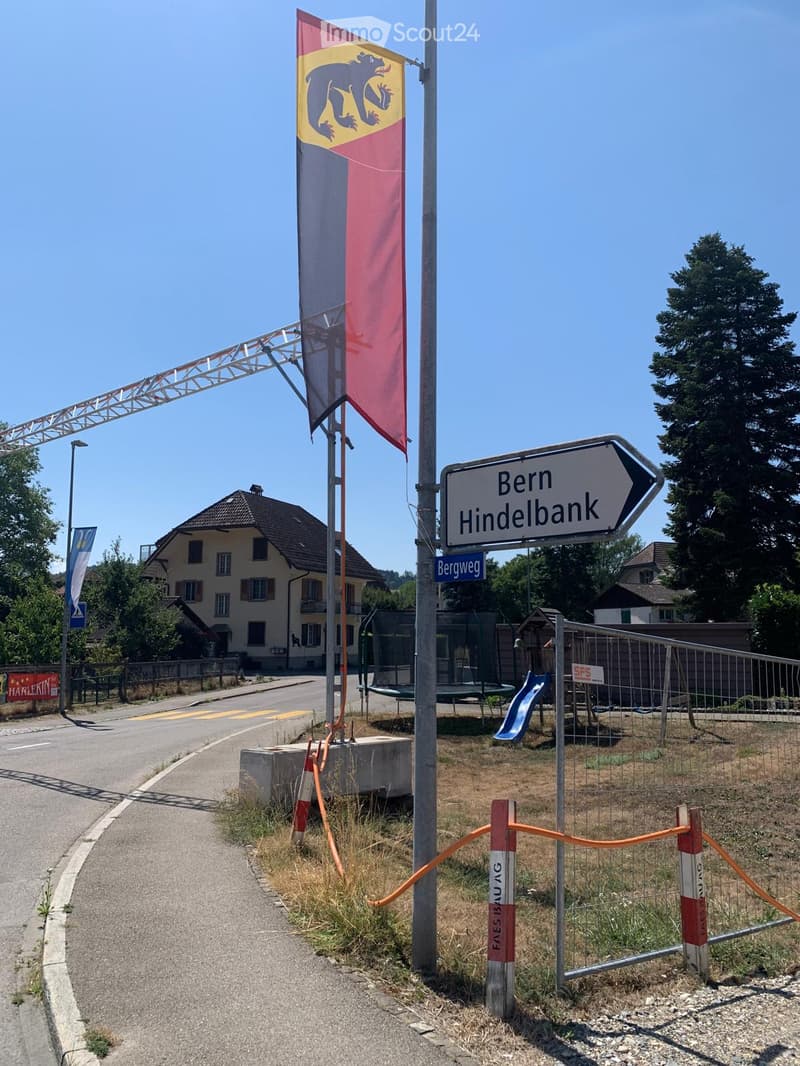 Outdoor area with a playground featuring a slide, a trampoline, and a fenced-off section. A road sign points to Bern and Hindelbank, with a colorful flag and a construction barrier visible.