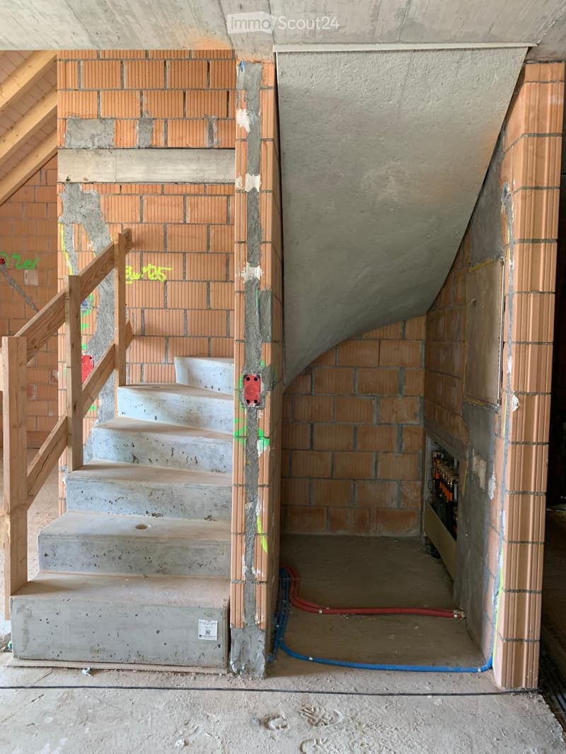 Staircase with concrete steps and wooden handrail, unfinished walls with exposed brick and concrete, visible electrical and plumbing installations.