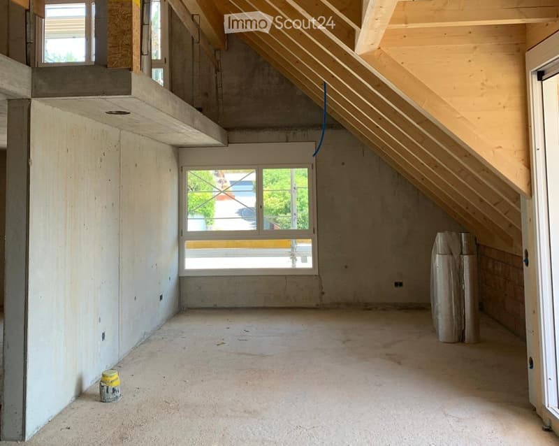 Empty attic space with exposed wooden beams, concrete walls, and a large window offering a view of the outside. The floor is unfinished concrete, and the room is completely unfurnished.