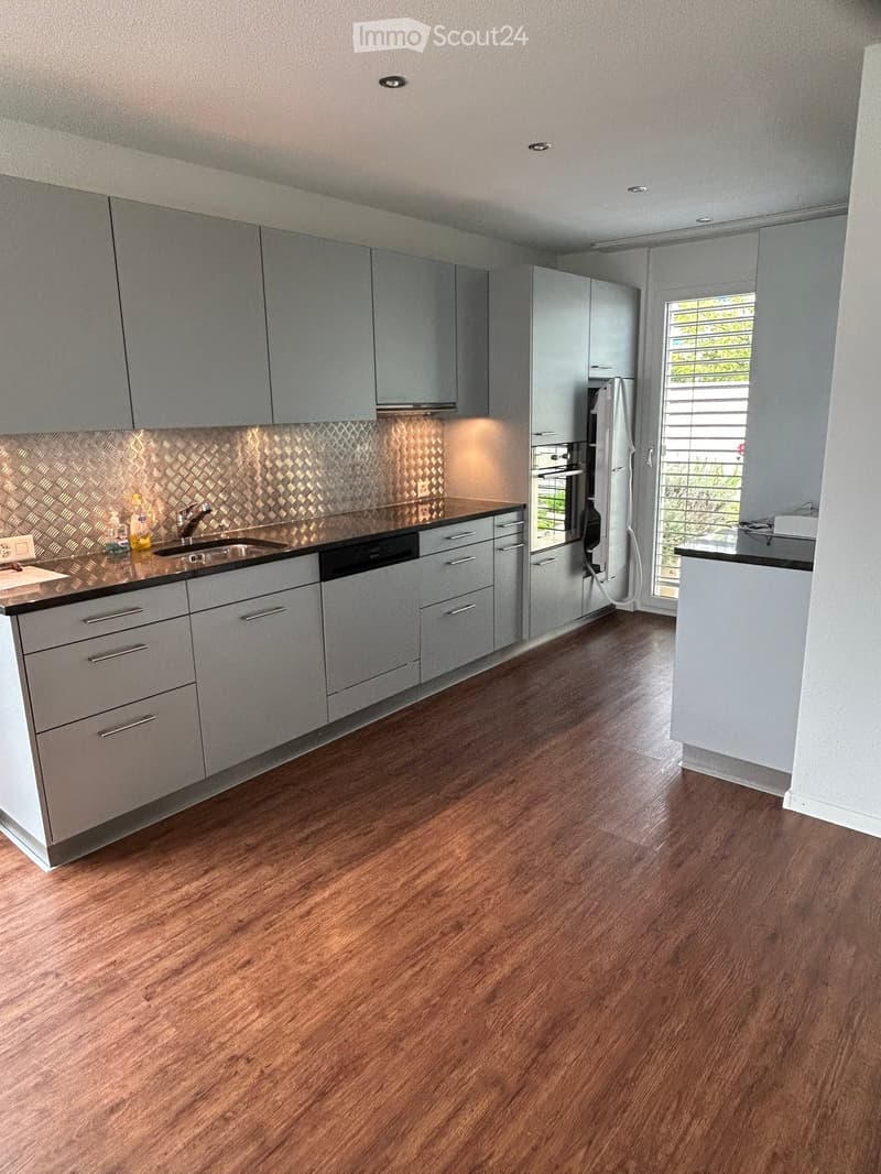 Modern kitchen with grey cabinets, wooden flooring, stainless steel appliances, and a patterned backsplash.