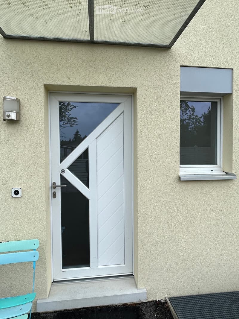 Exterior view of a building with a white door featuring a glass panel, a window with a white frame, and a light fixture mounted on the wall.