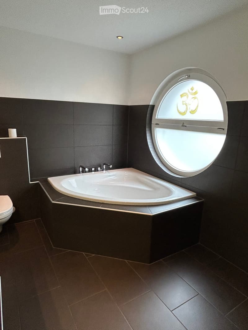 Modern bathroom with a freestanding bathtub, dark tiled walls, a round window with a decorative design, and a toilet visible in the background.