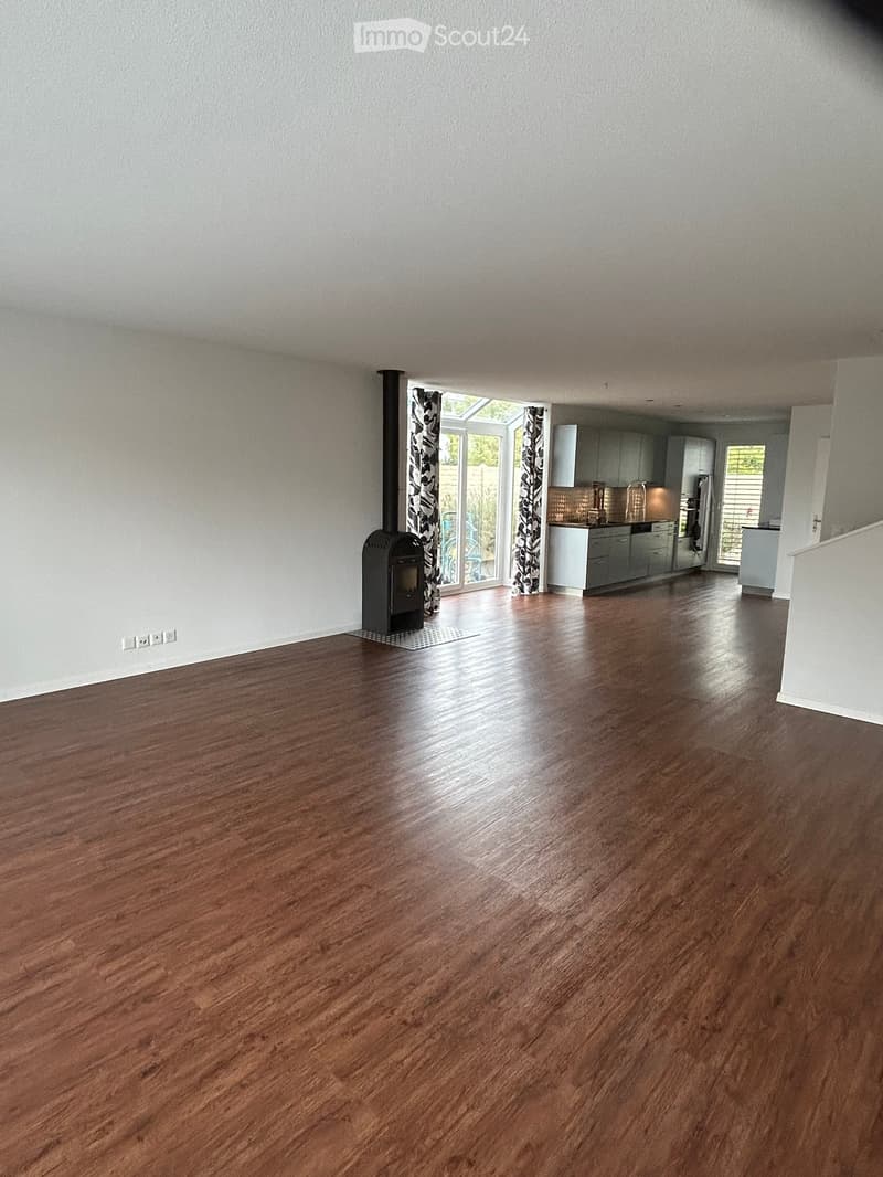 Empty room with wooden flooring, white walls, and a fireplace. The room is connected to a kitchen area with cabinets and appliances.