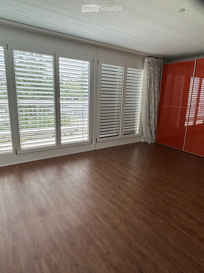 Empty room with wooden flooring, large windows with blinds, and a red closet door.