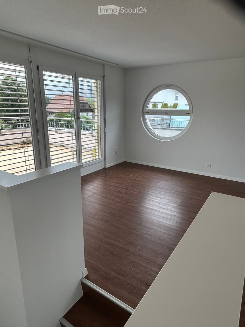 Empty room with wooden flooring, white walls, large window with blinds, and a round window overlooking a balcony area.