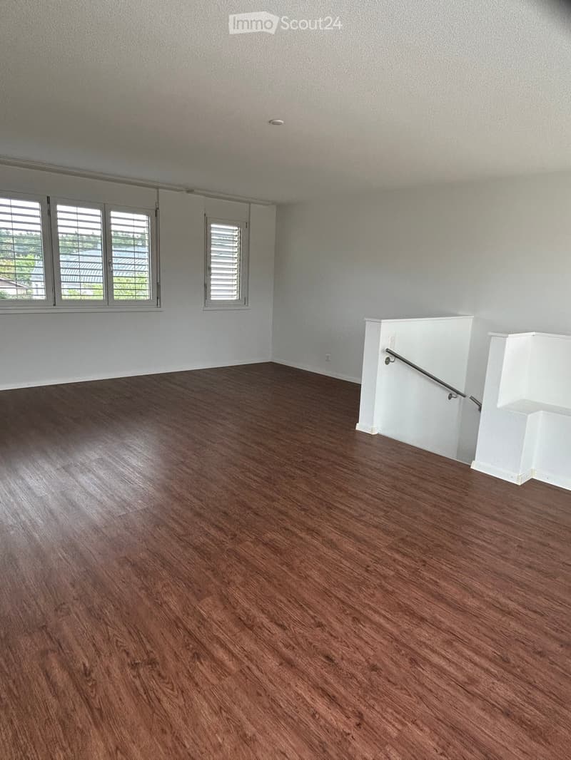 Empty room with wooden flooring, white walls, and large windows with blinds. A small white staircase with a handrail is visible on the right side.