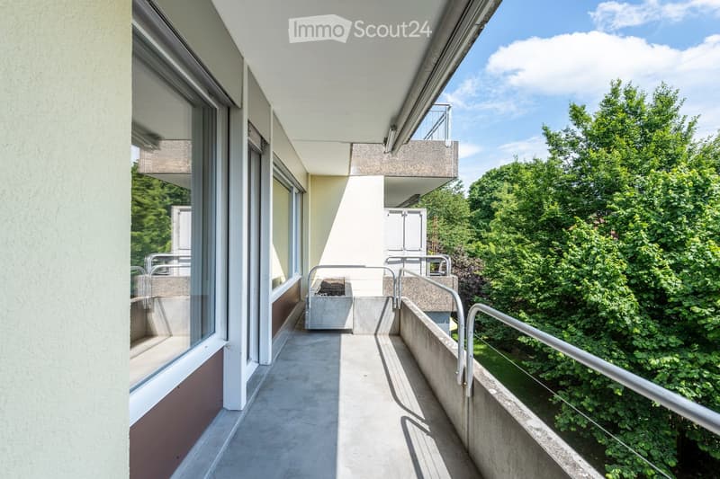 Modern balcony with concrete floor, metal railings, and large windows overlooking a green area with trees.