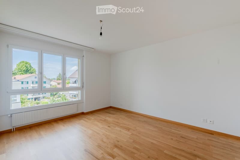 Empty room with light wooden flooring, white walls, large windows, radiator, and electrical outlets.