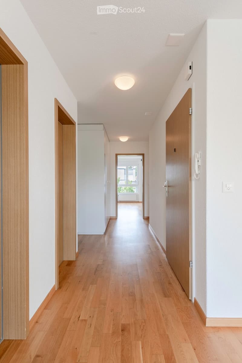 Bright hallway with wooden flooring, white walls, recessed lighting, and wooden doors on both sides. A small open area at the end leads to a room with a window and a radiator.