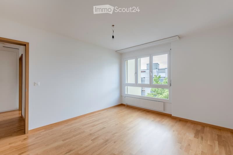 Empty room with light wooden flooring, white walls, and large windows letting in natural light. There is a radiator below the windows and a door on the left side.