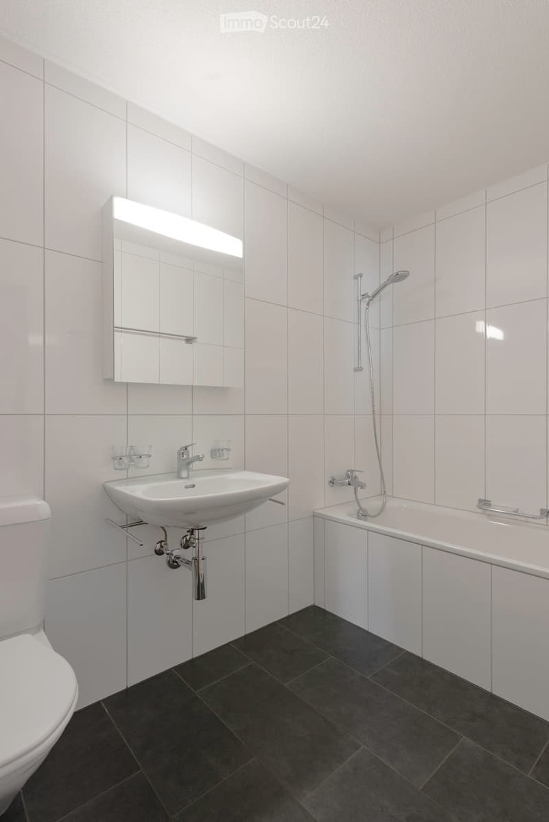 Modern bathroom with white tiles, a white sink, toilet, bathtub, and a mirror with built-in lighting. Dark tiled floor.