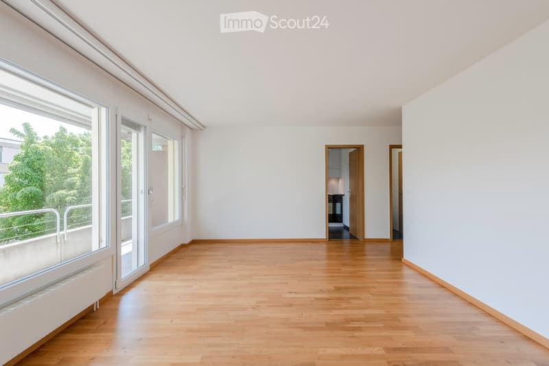 Empty room with wooden flooring, white walls, large windows, and sliding glass doors leading to a balcony.