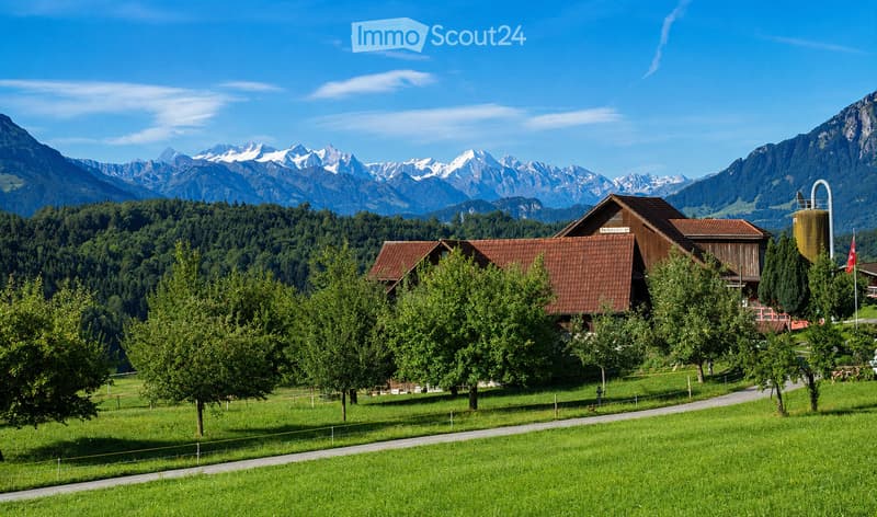 A picturesque rural scene with a traditional wooden house, surrounded by lush green trees and a well-maintained lawn, set against a backdrop of snow-capped mountains under a clear blue sky.