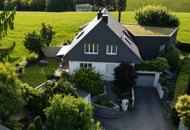 A modern two-story house with a dark grey roof and white walls, featuring a garage, garden, and surrounding greenery.