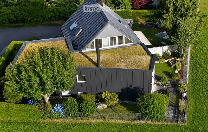 A modern two-story house with a grey exterior and a sloped roof, surrounded by a well-maintained garden with various plants and trees.