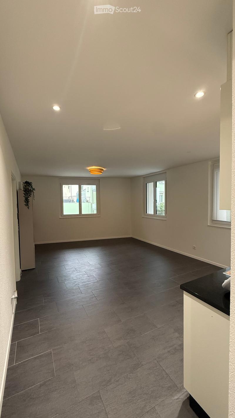 Empty room with light-colored walls, dark wooden flooring, and multiple windows allowing natural light. The room features recessed ceiling lights and a door leading to another area.