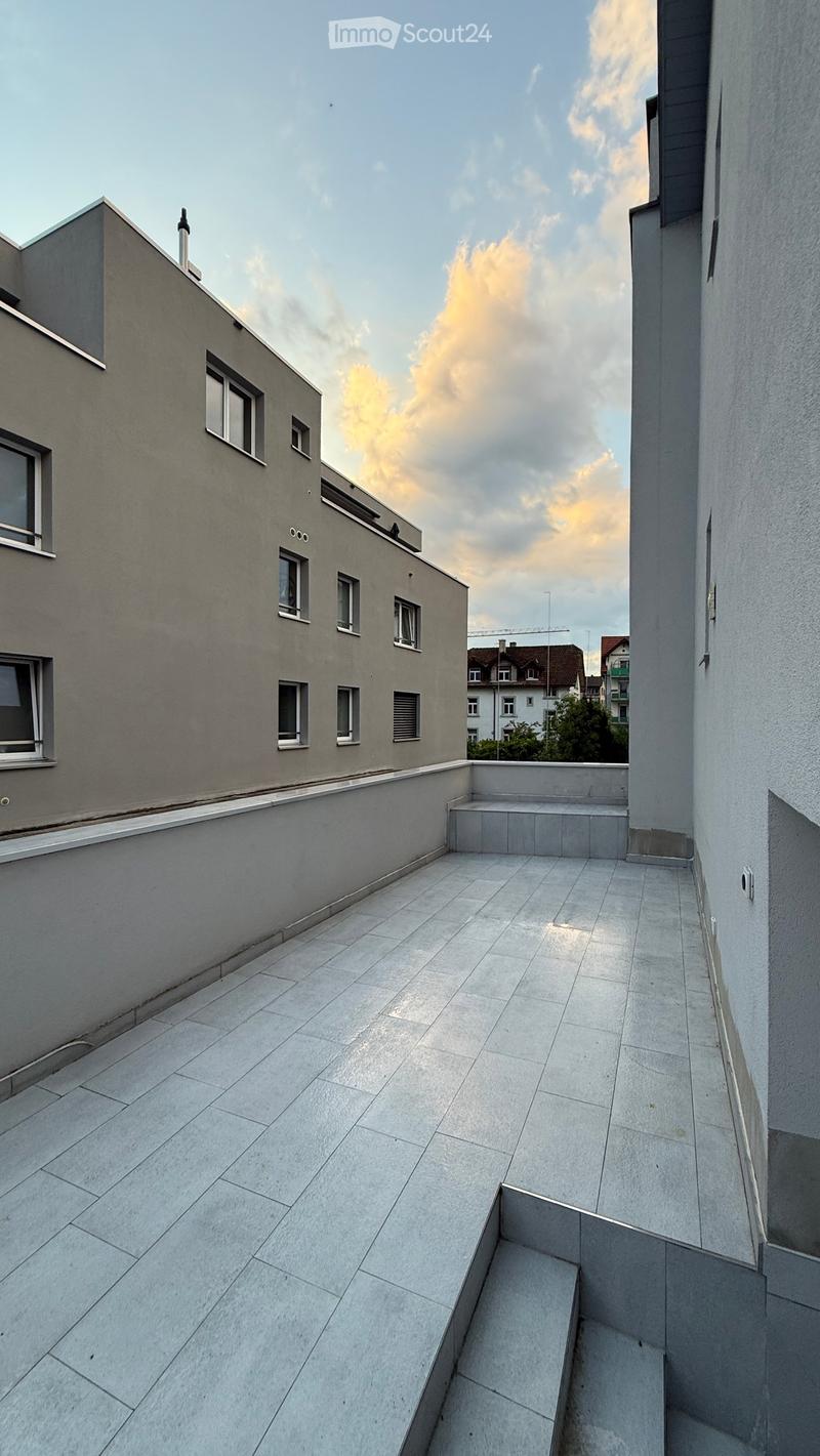 Modern rooftop terrace with grey tiled flooring, low walls, and steps leading to a raised platform, overlooking a cityscape with buildings and a partly cloudy sky.