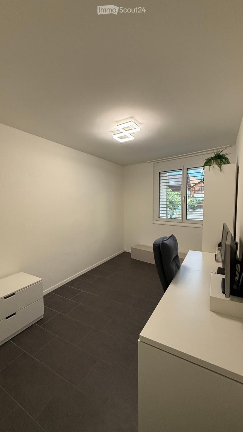 Modern office space with dark tiled floor, white walls, and a large window with blinds. Features a white desk with a screen, a black chair, and a white cabinet.