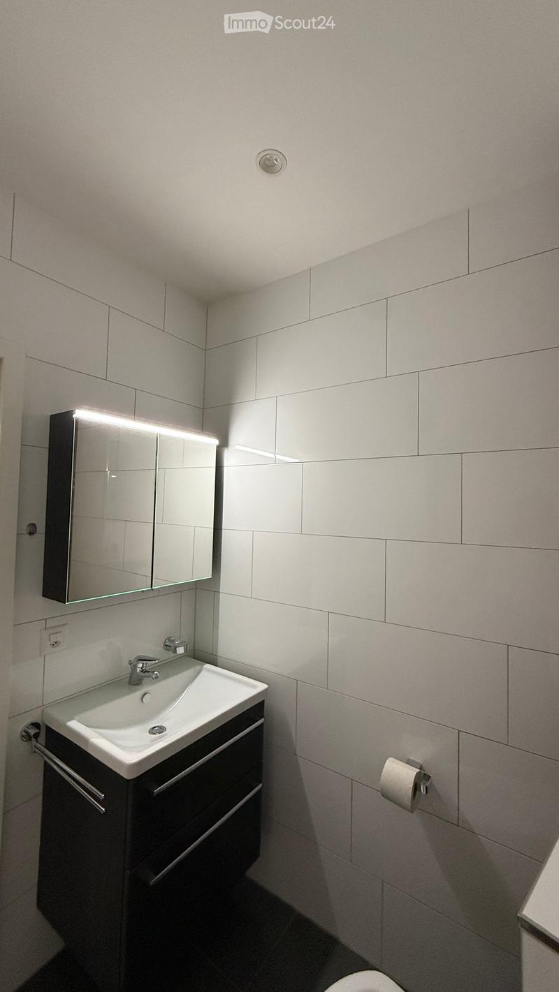 Modern bathroom with white tiles, a white sink with a chrome faucet, a mirrored cabinet with built-in lighting, and a dark wooden cabinet beneath the sink.