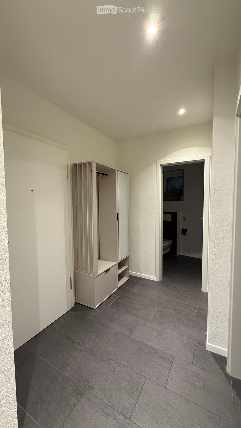 Empty hallway with grey tiled floor, white walls, and a built-in storage unit on the left. A doorway leads to a bathroom on the right.