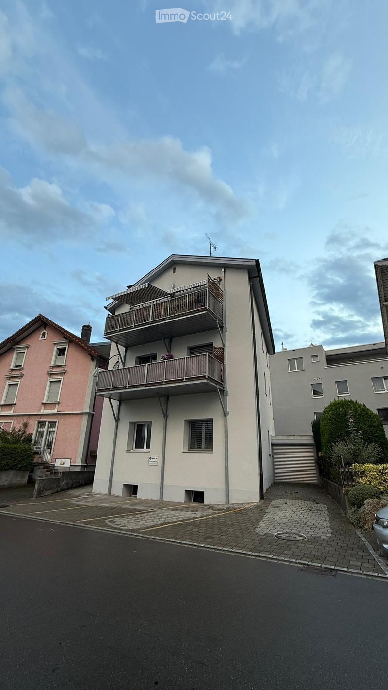 A two-story residential building with balconies, white exterior, and multiple windows. The building has a garage and is surrounded by other houses and greenery.