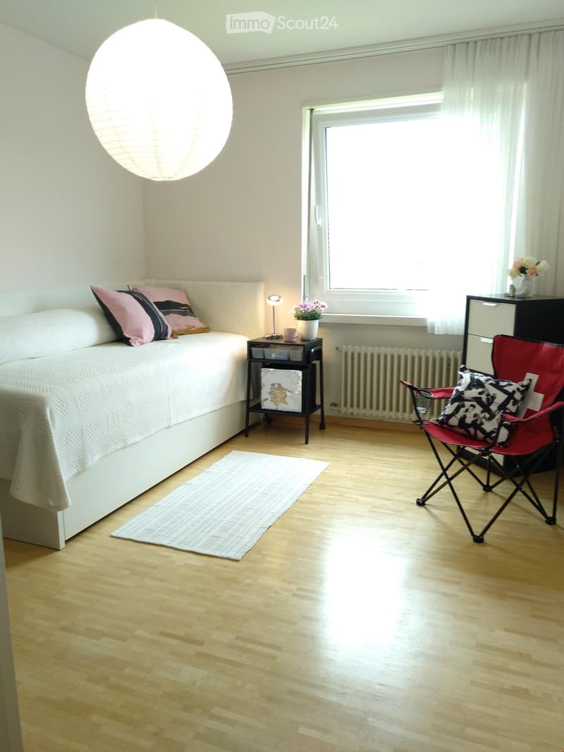 Modern bedroom with a white bed, pink and black pillows, a nightstand with a lamp, a red folding chair, and a large window with white curtains.