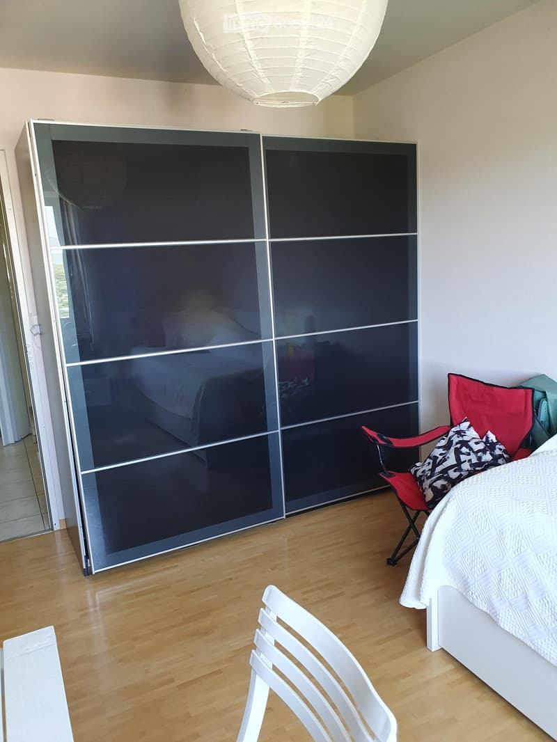 Modern bedroom with a white bed, red and black chair, white folding chair, and large dark wardrobe with mirrored doors.