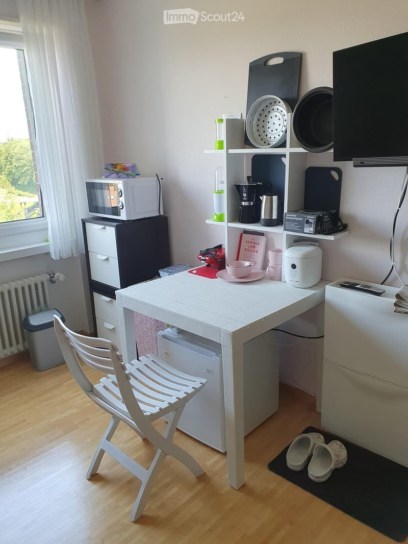 A small, well-lit room with a white wooden chair, a white table, a microwave, a shelf with various items, and a small refrigerator. The room also features a window with white curtains and a radiator below it.