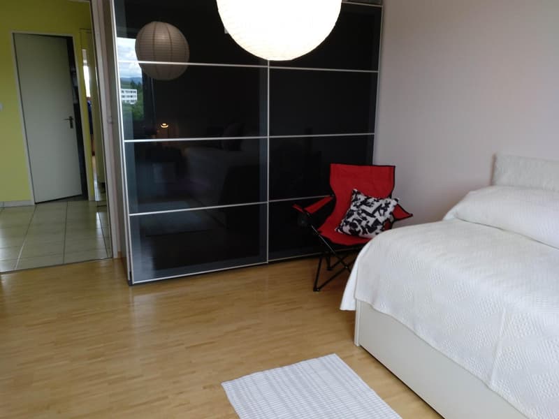 Modern bedroom with a white bed, red chair, and a large mirror reflecting another room. Wooden flooring and a white rug are visible.