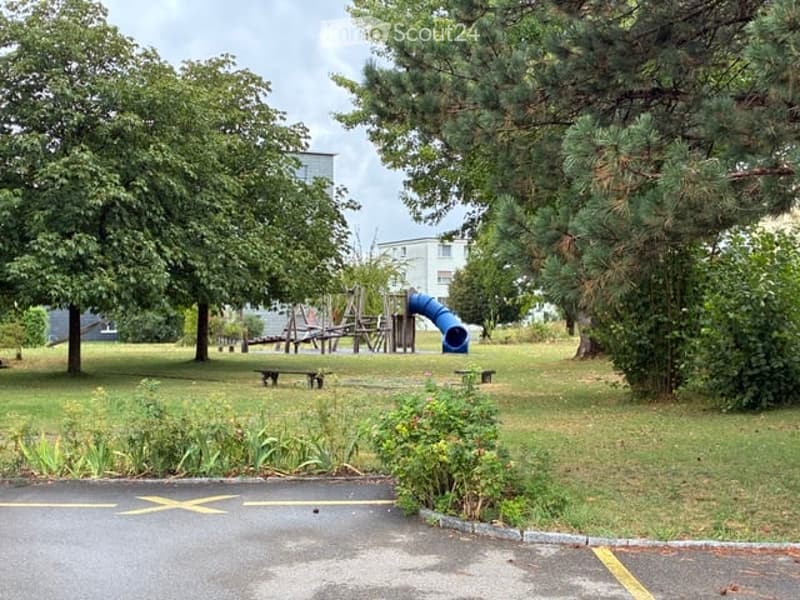A park-like outdoor area with a playground, benches, and trees, surrounded by a paved path.